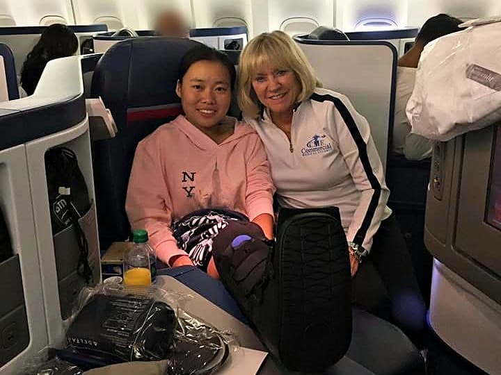 A CME escort in a branded jacket sitting beside a smiling patient whose leg is elevated in a walking boot, in a lie-flat airline seat